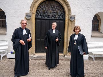 Ein Mann und zwei Frauen im Talar stehen vor einer Kirchentür.