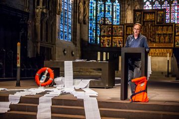 In der Dortmunder Stadtkirche werden am 20. Juni wieder Namen von gestorbenen Flüchtlingen verlesen. Foto: Stephan Schütze Ein Mann liest in einer Kirche Namen von verstorbenen Flüchtlingen vor. Daneben liegen lange Listen mit den Namen darauf.